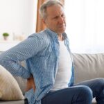 Man holding his lower back in pain, sitting on a couch.